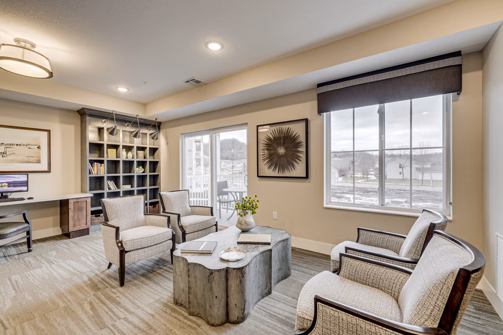 Bright communal sitting area with four upholstered armchairs around a sculpted coffee table, bookshelves, a desk, and large windows.