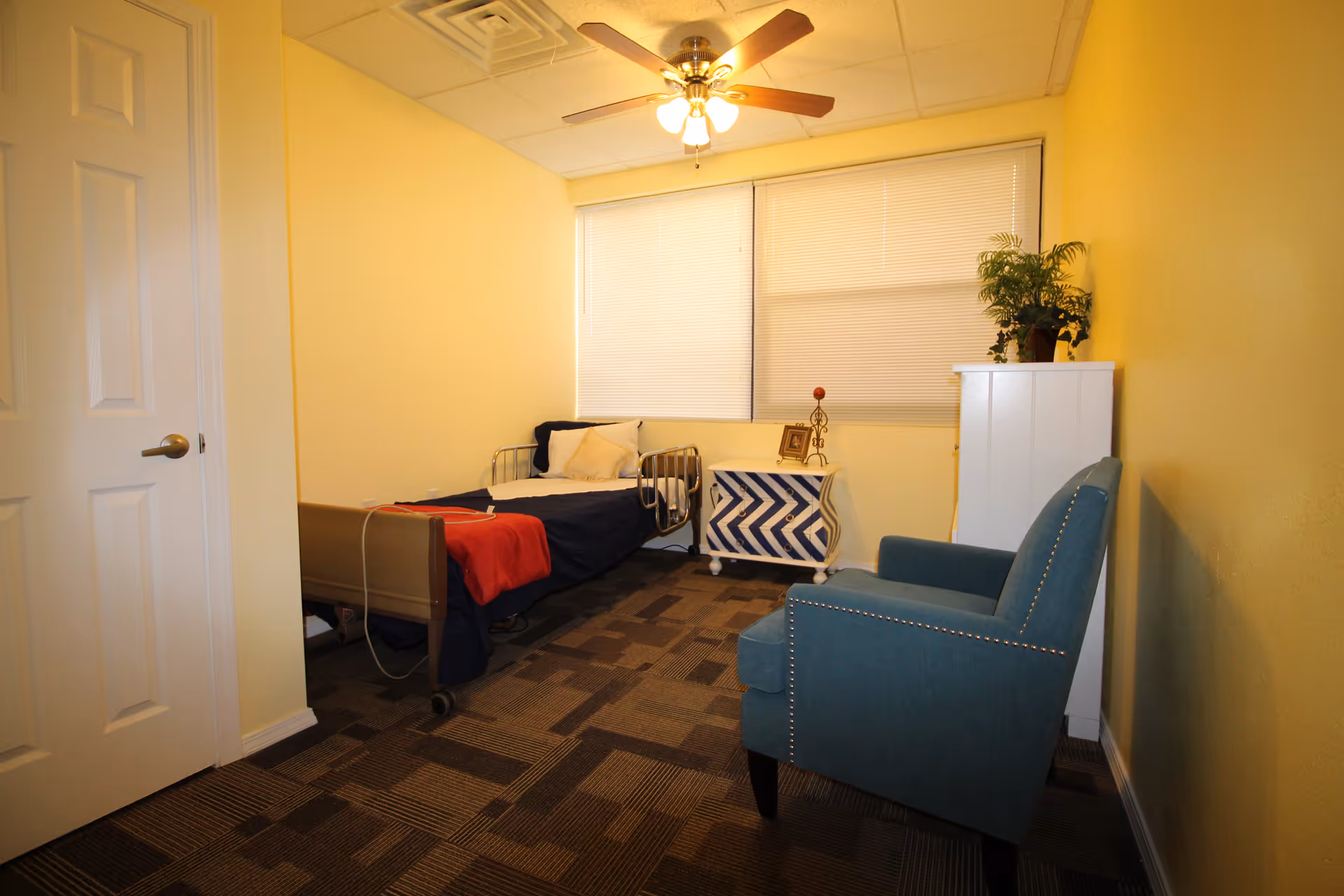 A small bedroom with yellow walls and a patterned carpet floor. The room contains a single bed with metal side rails, dressed with dark blue bedding and a red blanket. Next to the bed is a white nightstand with a blue zigzag pattern, holding a small framed picture and a decorative item. A blue upholstered armchair is positioned against the right wall, and a white cabinet with a potted plant on top is also visible. The ceiling has a fan with lights, and a large window with closed blinds is on the back wall.