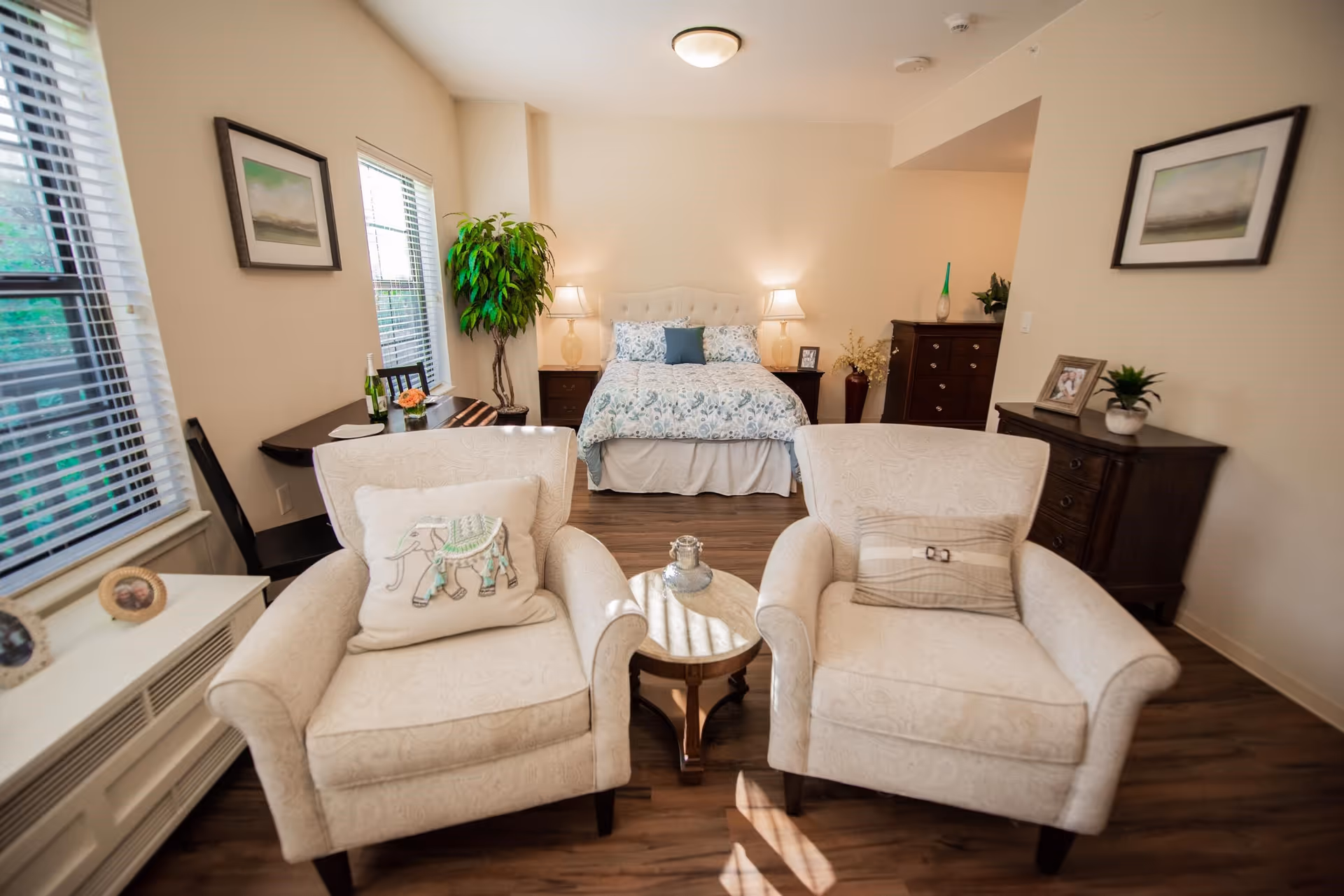 A cozy senior living bedroom with a bed in the background, two beige armchairs with decorative pillows in the foreground, a small round table between the chairs, wooden flooring, framed pictures on the walls, a potted plant near the window, and a dresser with decorative items.