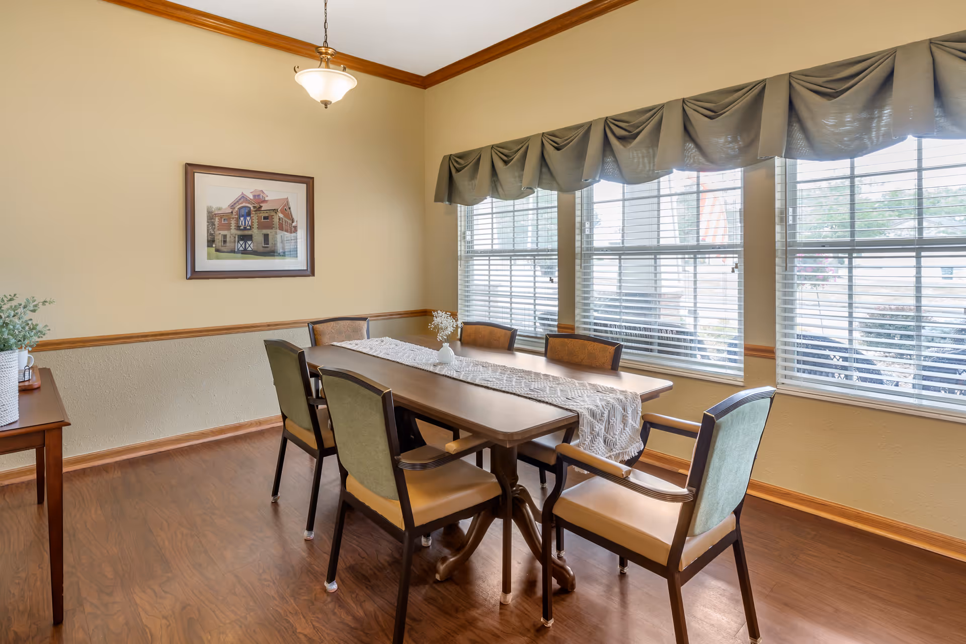 A dining room with a wooden table covered by a white lace runner and six chairs around it. The room has large windows with blinds and green valances, a wooden floor, beige walls with a chair rail, a framed picture on the wall, and a small table with a plant on the left side.