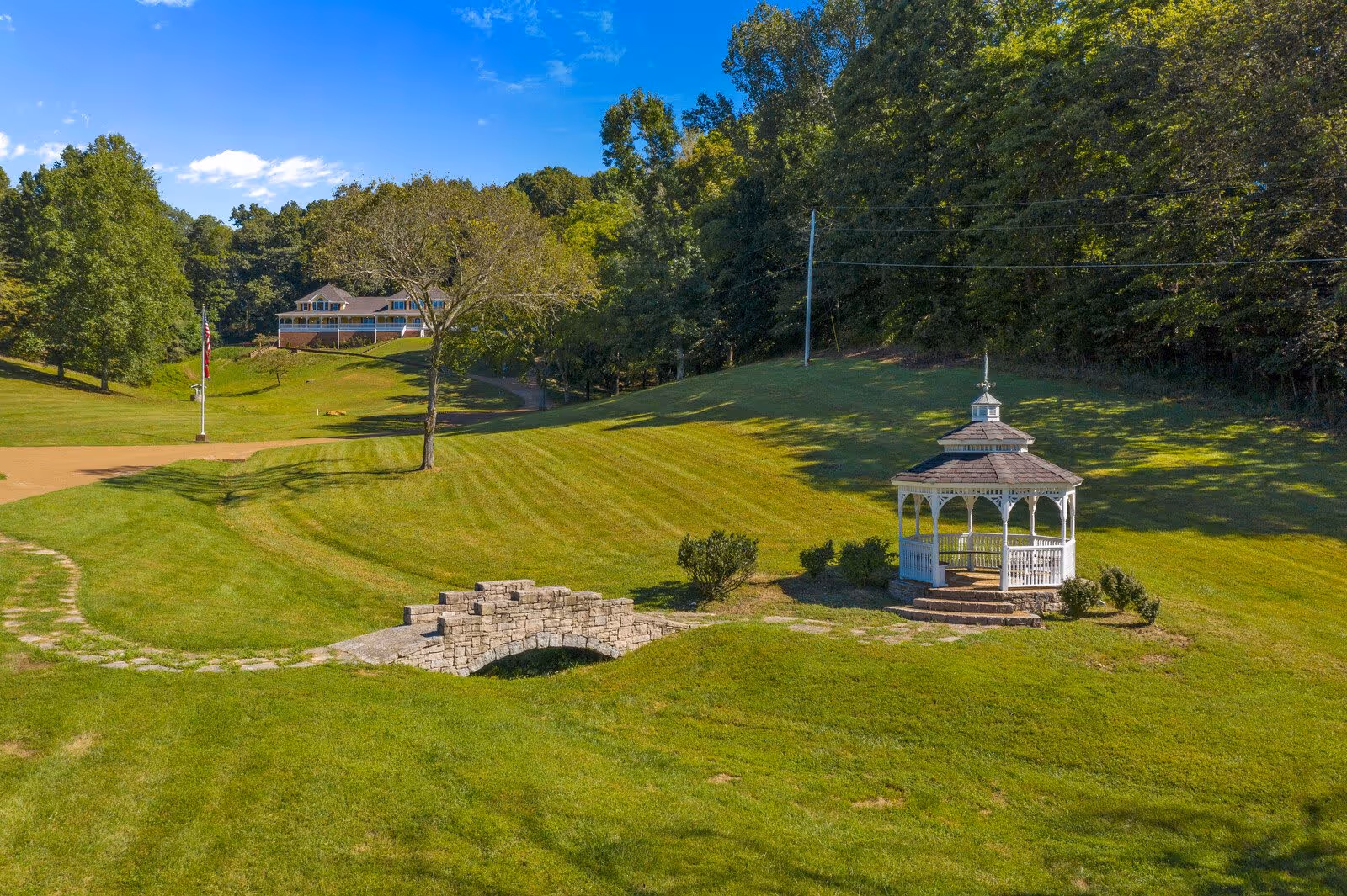 A scenic outdoor area at New Hope Senior Living of Hendersonville featuring a well-maintained grassy lawn, a small stone bridge over a shallow ditch, a white gazebo with a shingled roof, and a large house in the background surrounded by trees under a clear blue sky.