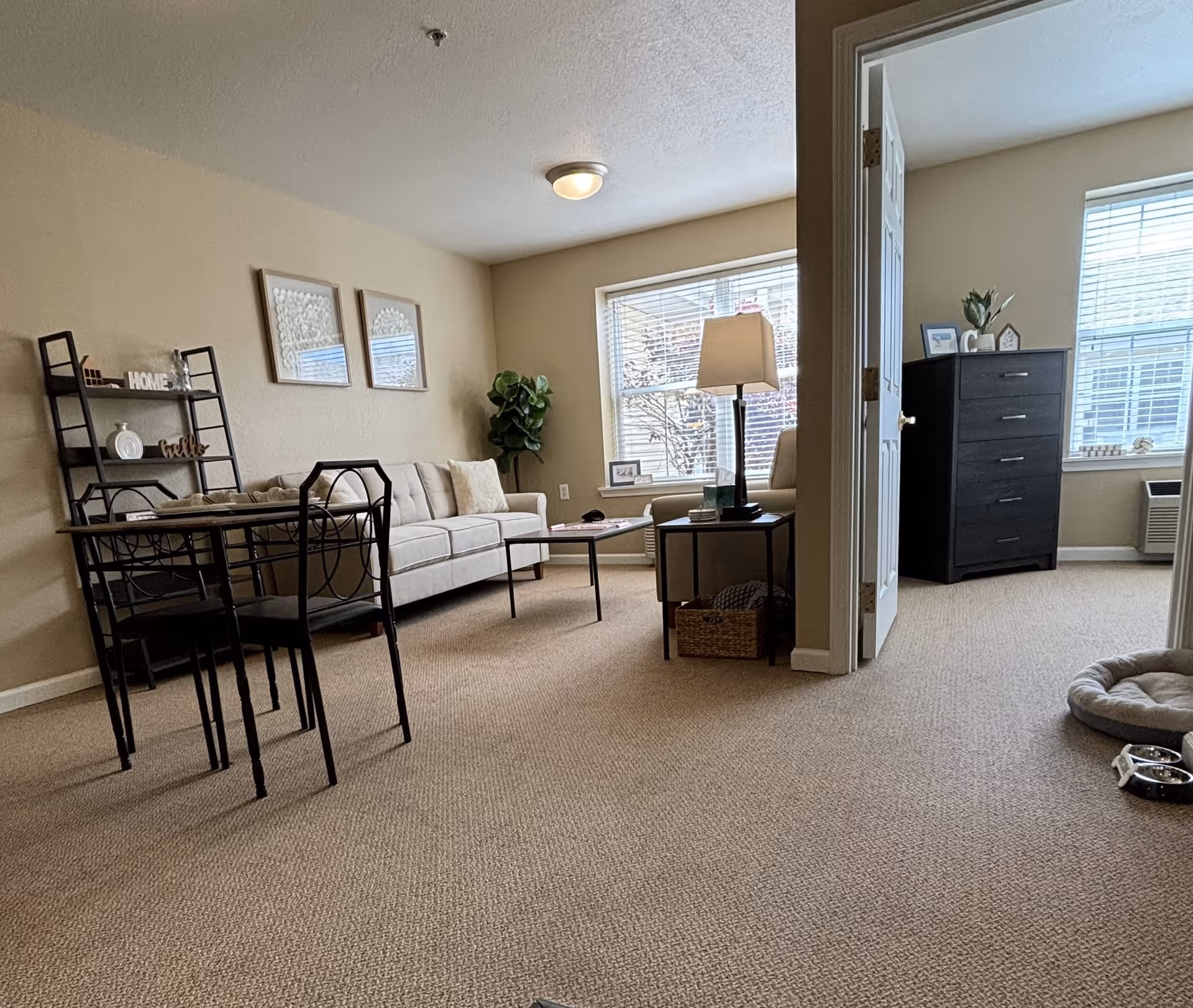 A cozy living area in a senior living facility with a beige sofa, a black metal dining table with chairs, a floor lamp, and a small coffee table. There is a doorway leading to a bedroom with a black dresser and windows with blinds. The room is carpeted and decorated with framed art and a potted plant.