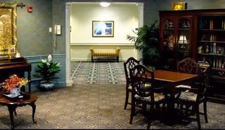 Carpeted communal sitting area with a wooden table and chairs, a bookshelf, potted plants, and a bench visible in the hallway.