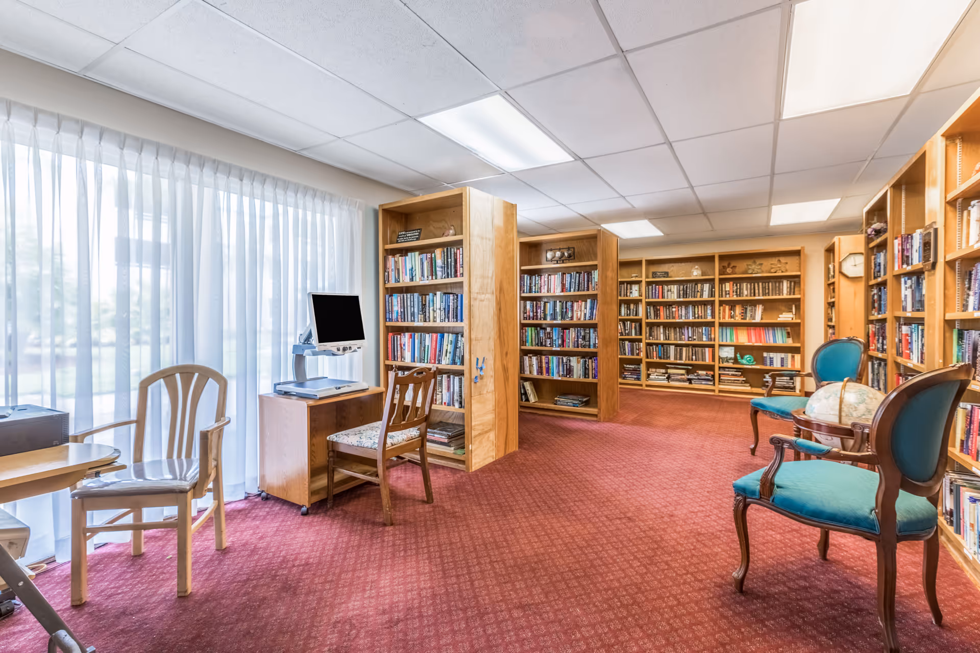 A cozy library room with red carpet flooring, wooden bookshelves filled with books, a computer on a small wooden desk with a chair, and two teal upholstered chairs with wooden frames. Large windows with sheer white curtains allow natural light to fill the space.