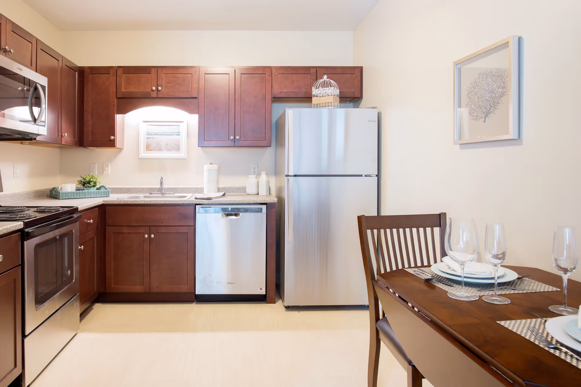 A modern kitchen with wooden cabinets, stainless steel appliances including a microwave, oven, dishwasher, and refrigerator. There is a double sink under a cabinet with a framed picture above it. To the right, a wooden dining table is set with plates, napkins, and wine glasses, and a framed artwork hangs on the wall.