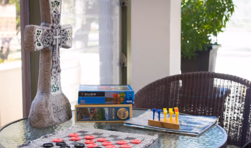 A glass-top round table with a stone cross decoration inscribed with the word 'Believe', two stacked puzzle boxes, a wooden peg game with colorful pegs, and a checkers board with red and black pieces. The table is situated near a window with natural light and a wicker chair is visible in the background along with a potted plant.