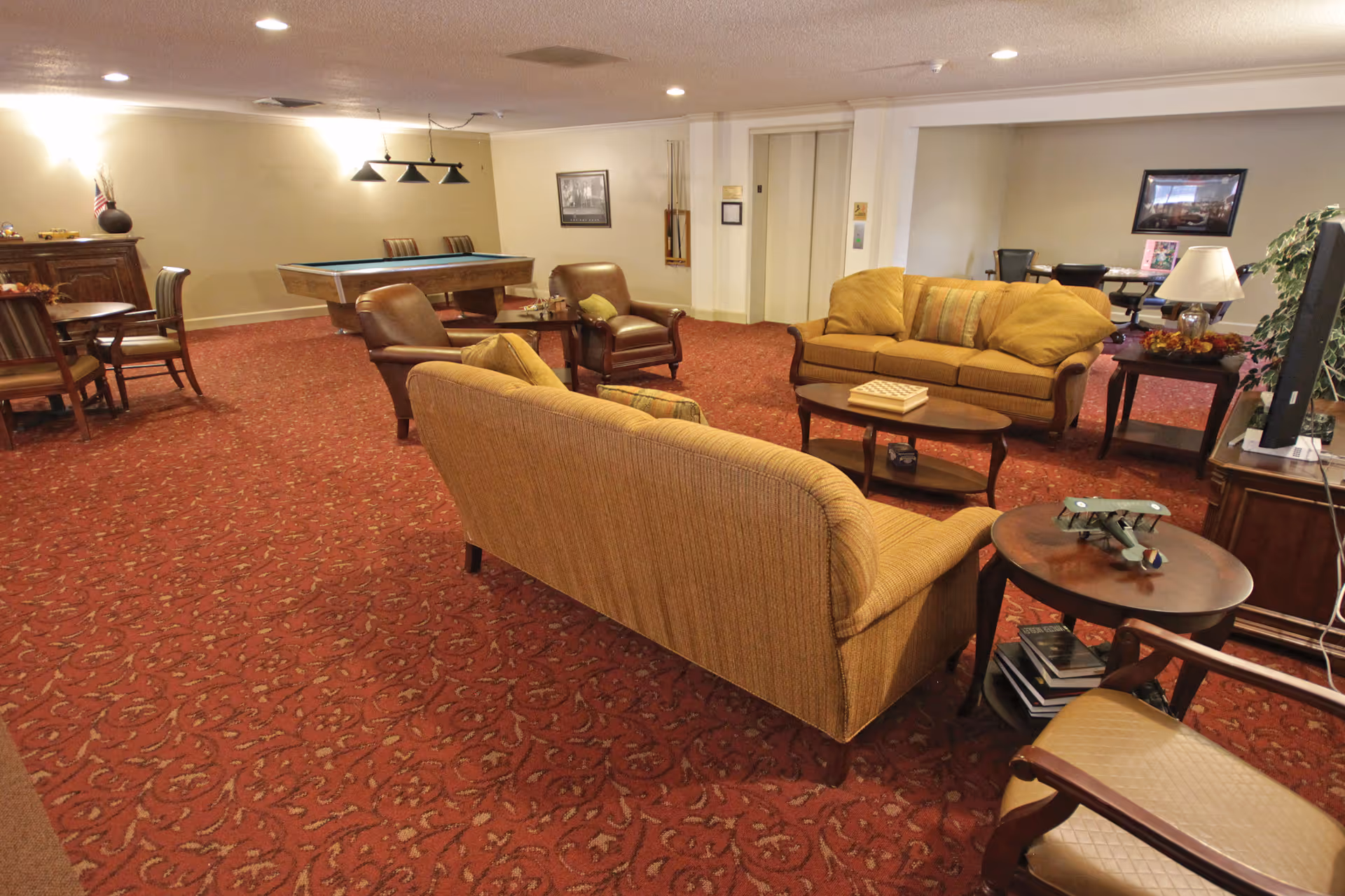 Community lounge with sofas, armchairs, side tables, and a pool table on red patterned carpet.