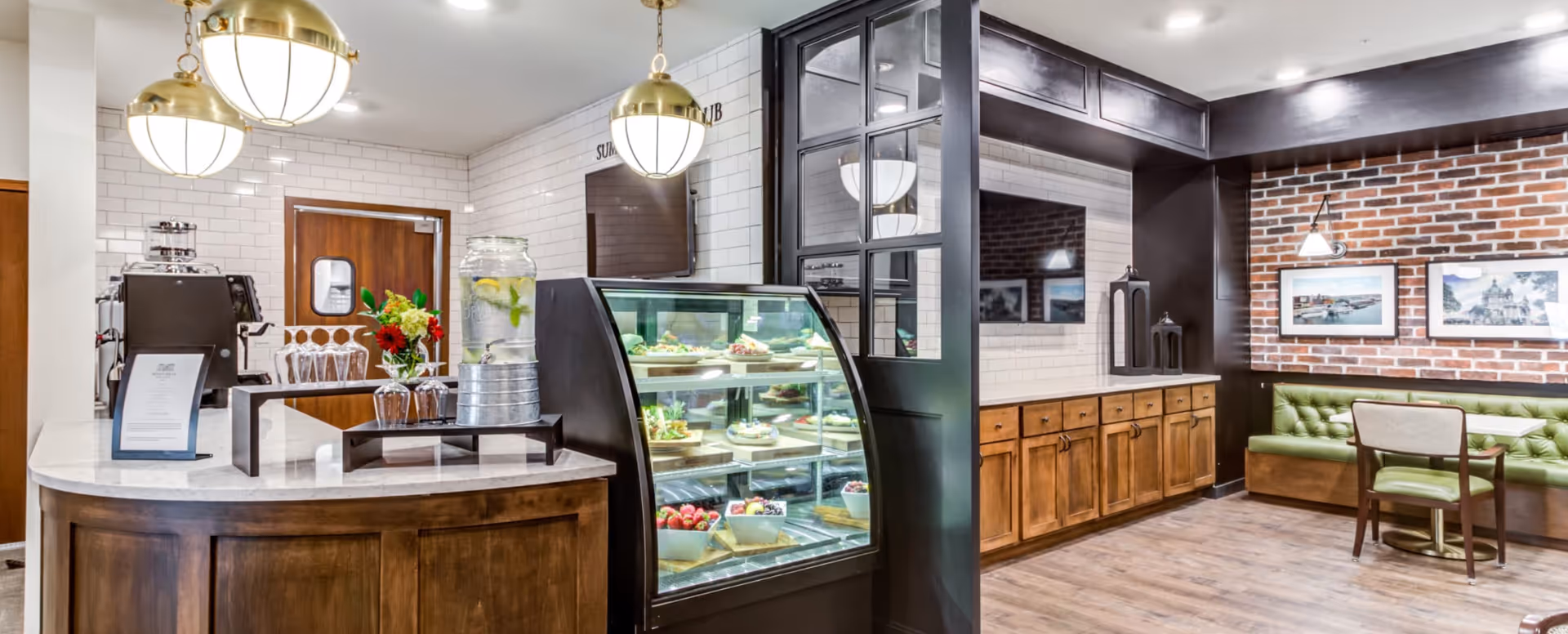 Bright senior-living café and dining area with a glass pastry display, service counter, and banquette seating by a brick accent wall.
