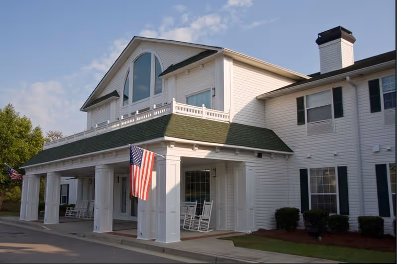 Front entrance of a white senior living facility with columns, rocking chairs on the porch, and an American flag.