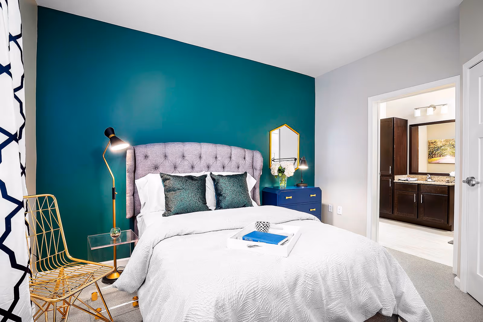 A modern bedroom with a tufted gray headboard against a teal accent wall, a white bedspread, two green decorative pillows, a gold wire chair, a floor lamp, and a blue nightstand with a small mirror and flowers. An open door reveals a bathroom with dark wood cabinets and a large mirror.