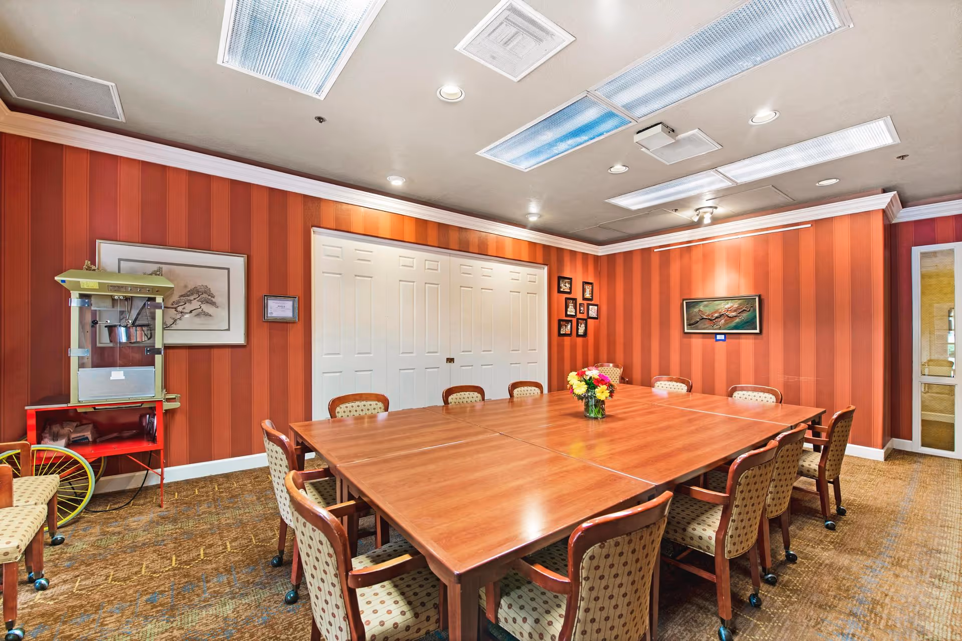 A meeting or dining room with a large rectangular wooden table surrounded by twelve cushioned chairs with patterned upholstery. The walls are painted with vertical red stripes and decorated with framed artwork and photos. There is a popcorn machine on a red cart in the corner and a vase with colorful flowers on the table. The ceiling has recessed lighting and large fluorescent light panels.