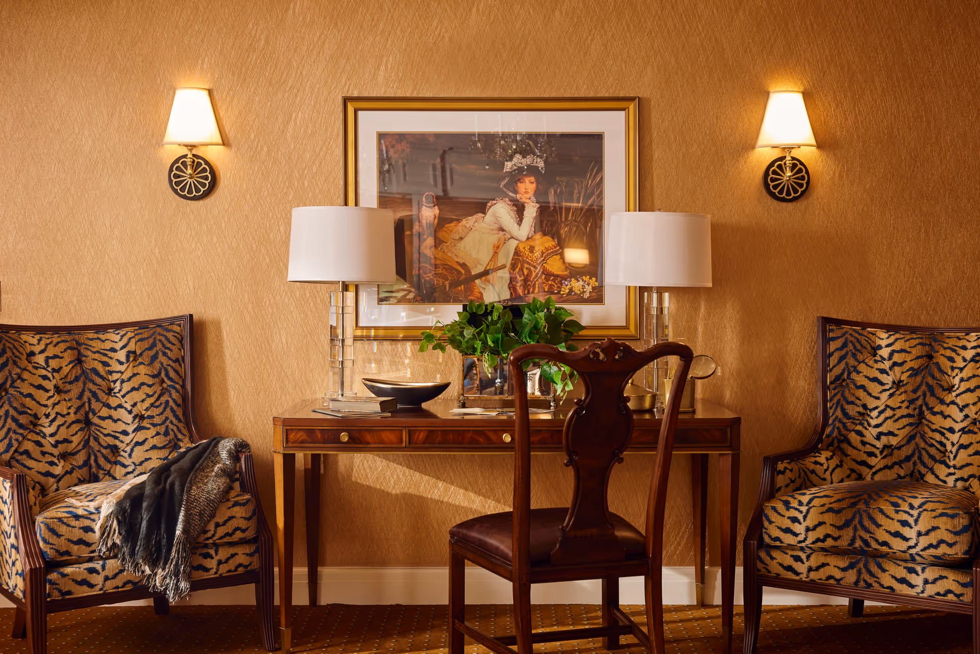 A cozy interior space featuring a wooden desk with two clear glass lamps and a green plant on top. Behind the desk is a framed painting of a woman in historical attire. On either side of the desk are two armchairs with tiger-striped upholstery, one draped with a dark throw blanket. The walls are covered in a textured beige wallpaper, and two wall sconces with lampshades provide warm lighting.