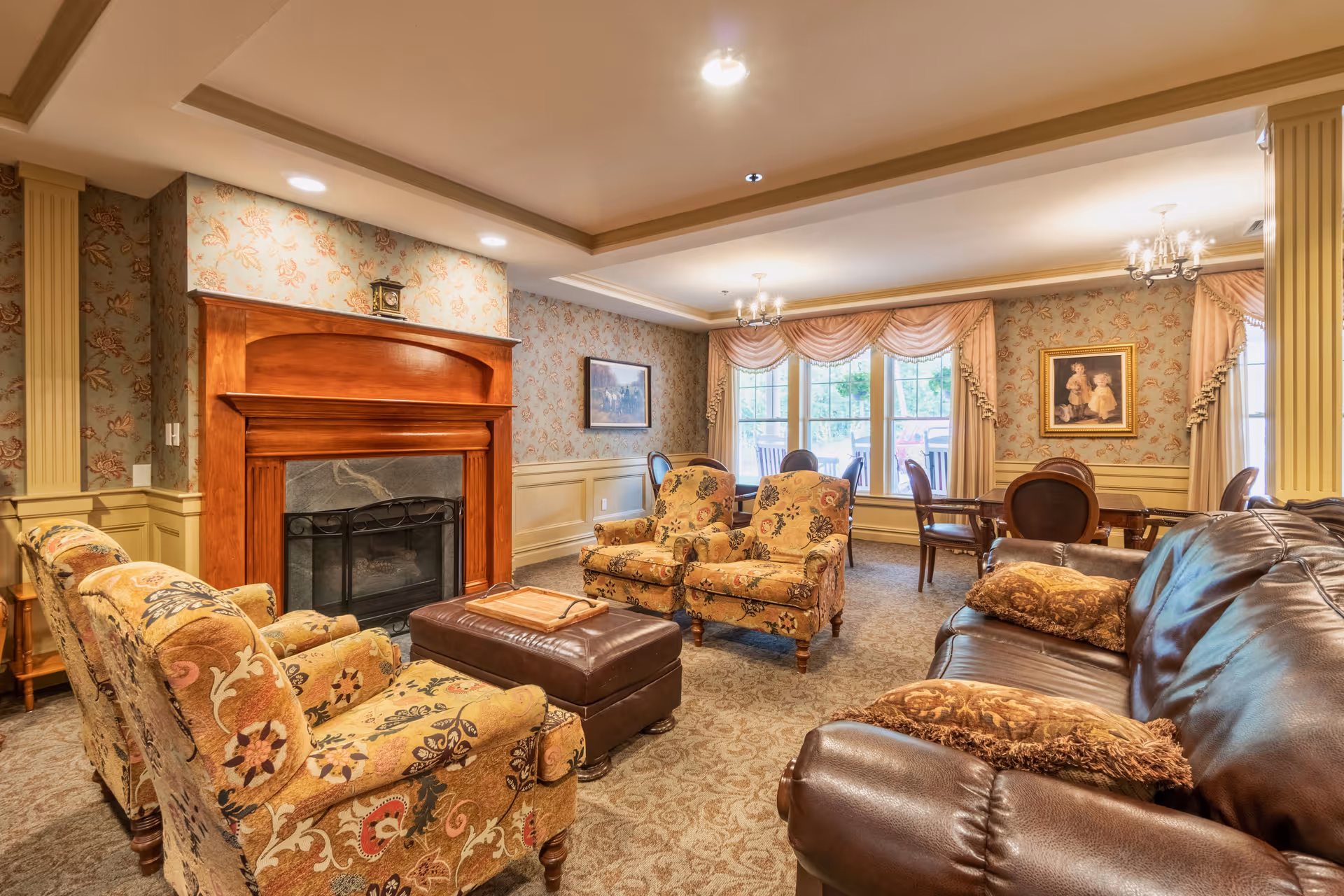 Cozy lounge with patterned upholstered chairs, a leather sofa, a fireplace, and a dining table by large windows.