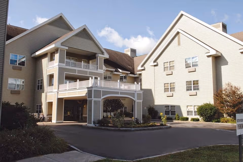 Front exterior of a multi-story beige senior living building with a covered porte-cochere, balconies, and a circular driveway.
