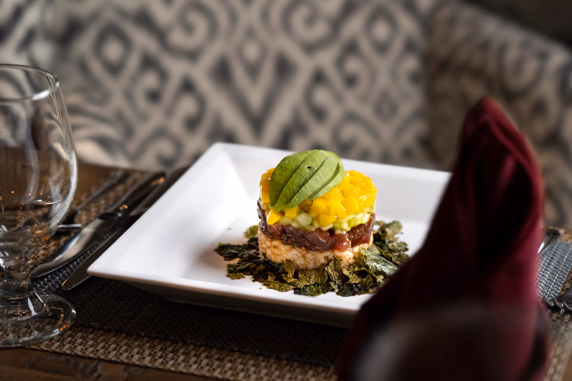 A layered seafood tartare topped with sliced avocado and mango on a square white plate set on a dining table.