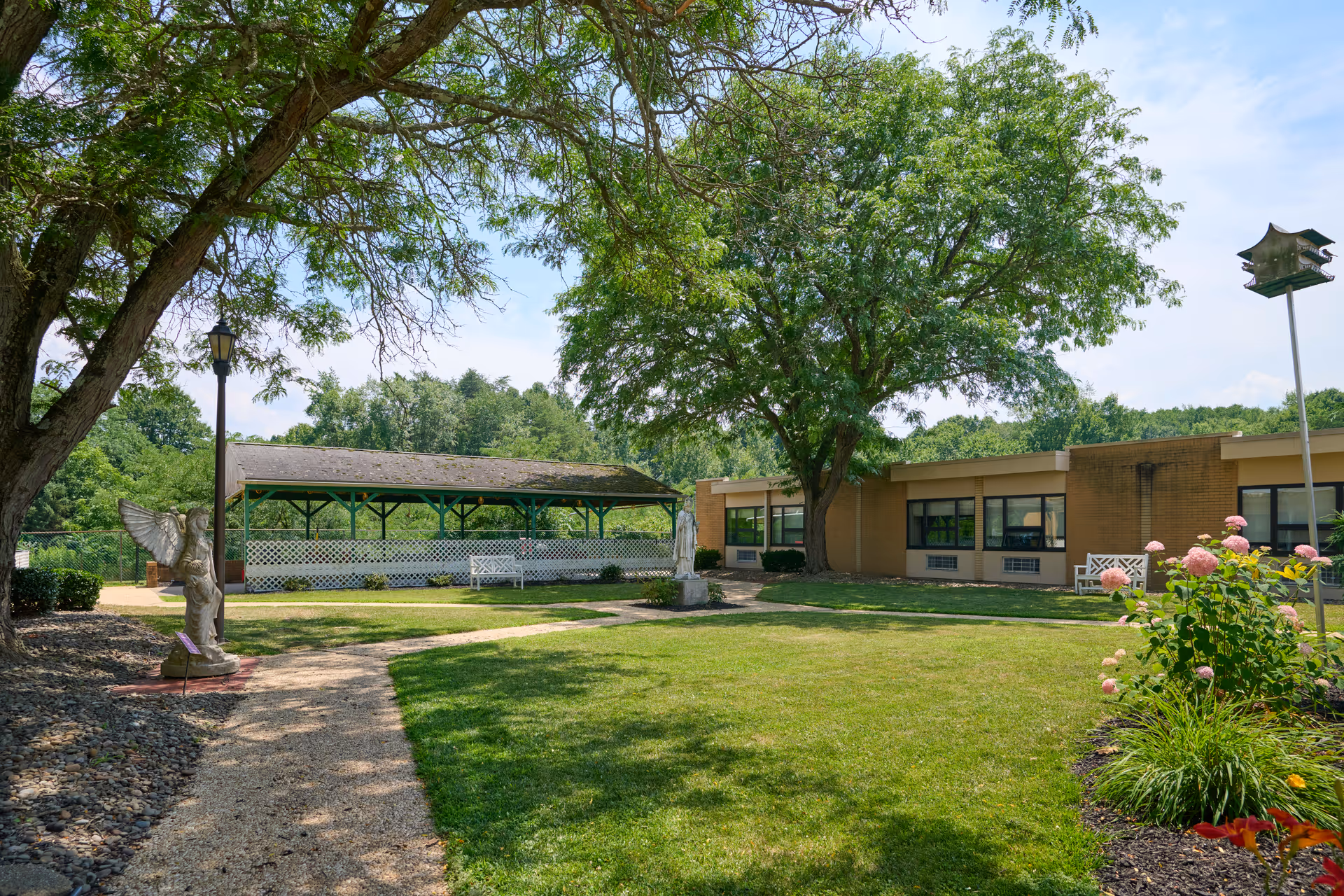 Outdoor garden area at Lakewood Rehabilitation and Healthcare Center featuring a well-maintained lawn, trees providing shade, flower beds with blooming flowers, a covered seating area with benches, statues, and a birdhouse on a tall pole.