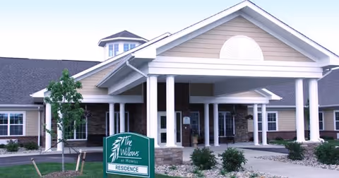 Exterior view of The Willows At Howell senior living residence showing the front entrance with a covered driveway supported by white columns, landscaped bushes, and a green sign with the facility name.