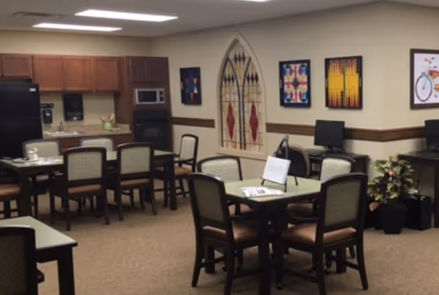 Communal dining/activity room with multiple tables and chairs, a kitchenette along the back wall, computers and wall art.
