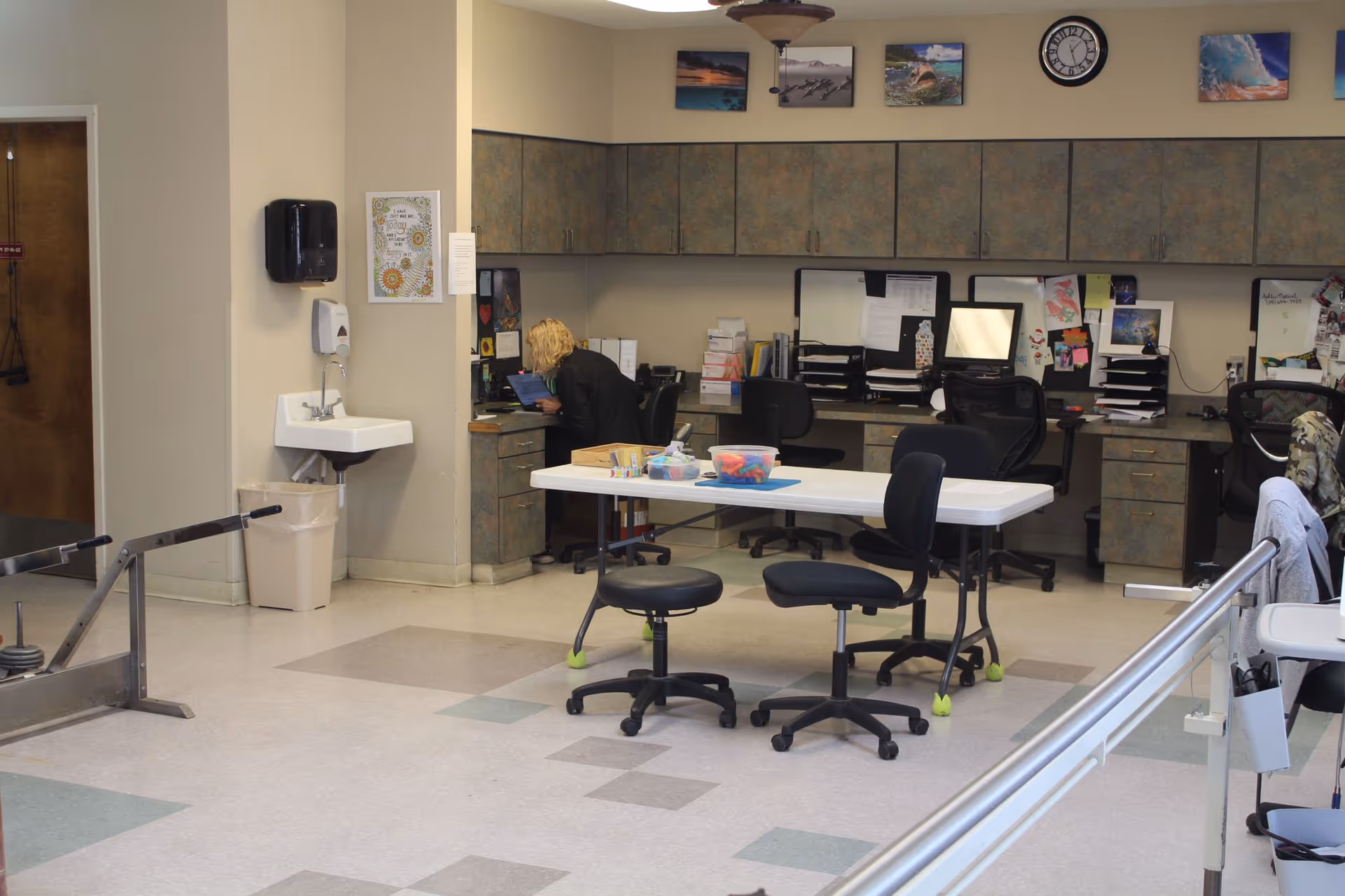 Interior of a rehabilitation/therapy room showing a staff member at a desk, a central folding table with chairs, cabinets along the back wall, and parallel walking bars in the foreground.