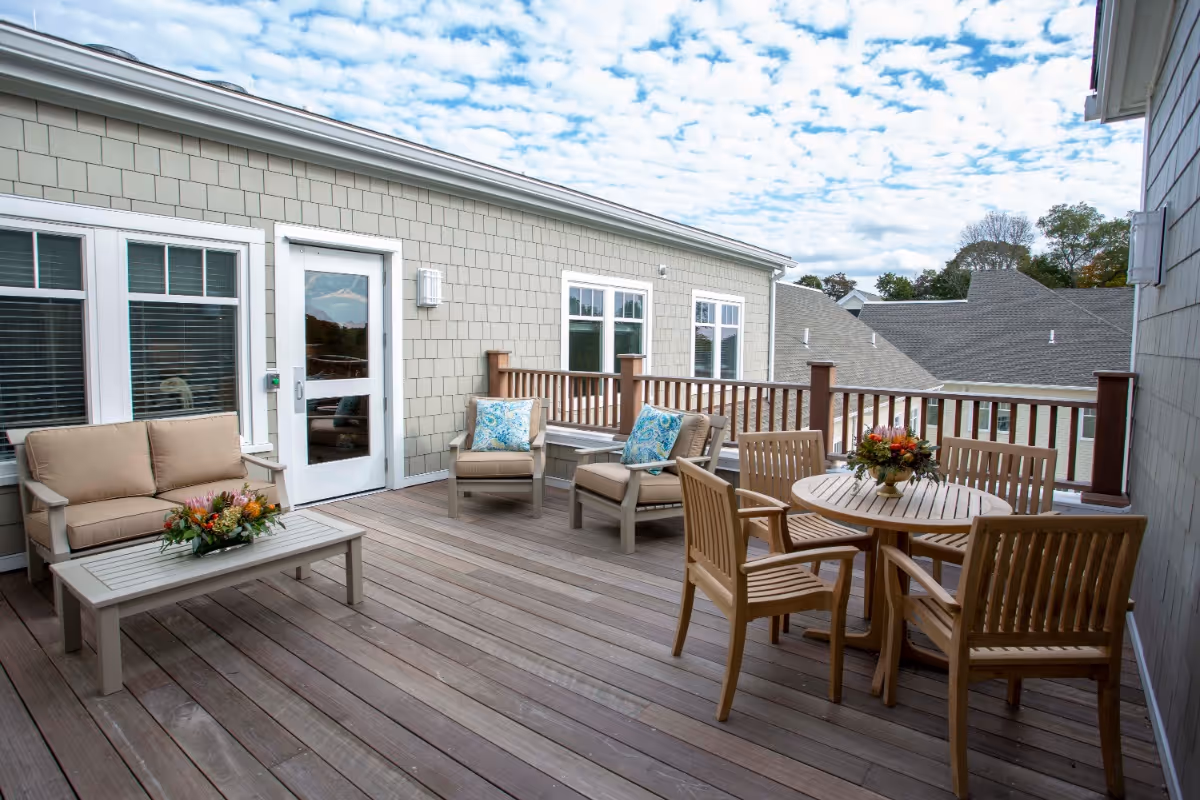 Outdoor patio area with wooden deck flooring, featuring a seating arrangement including a beige cushioned loveseat, two cushioned armchairs with blue patterned pillows, a coffee table with a floral centerpiece, and a round wooden dining table with four chairs and a floral centerpiece. The patio is enclosed by a wooden railing and is adjacent to a building with light gray siding and white-framed windows and door. The sky is partly cloudy.