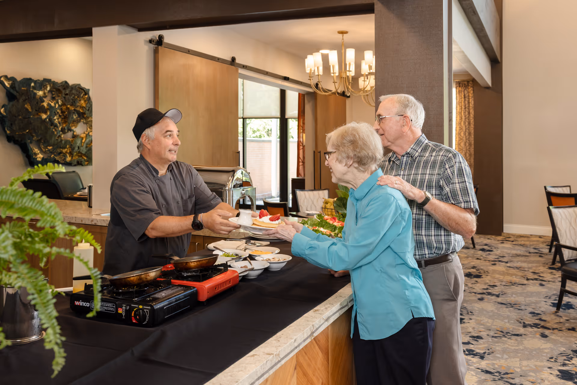 A chef wearing a black cap and uniform hands a plate of food to an elderly woman in a blue shirt, while an elderly man in a plaid shirt stands beside her with his hand on her shoulder. They are at a serving counter in a dining area with tables and chairs in the background.