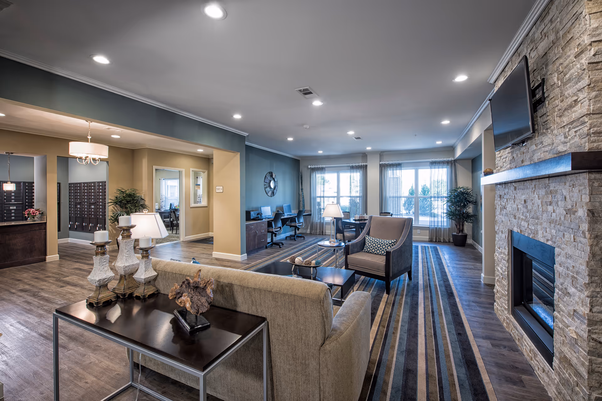 A spacious and well-lit living room area in a senior living facility featuring a stone fireplace with a mounted TV above it, a beige sofa with a side table holding decorative lamps and a sculpture, a comfortable armchair with a patterned pillow, a striped area rug, and a work area with desks and chairs near large windows with sheer curtains allowing natural light to fill the room.