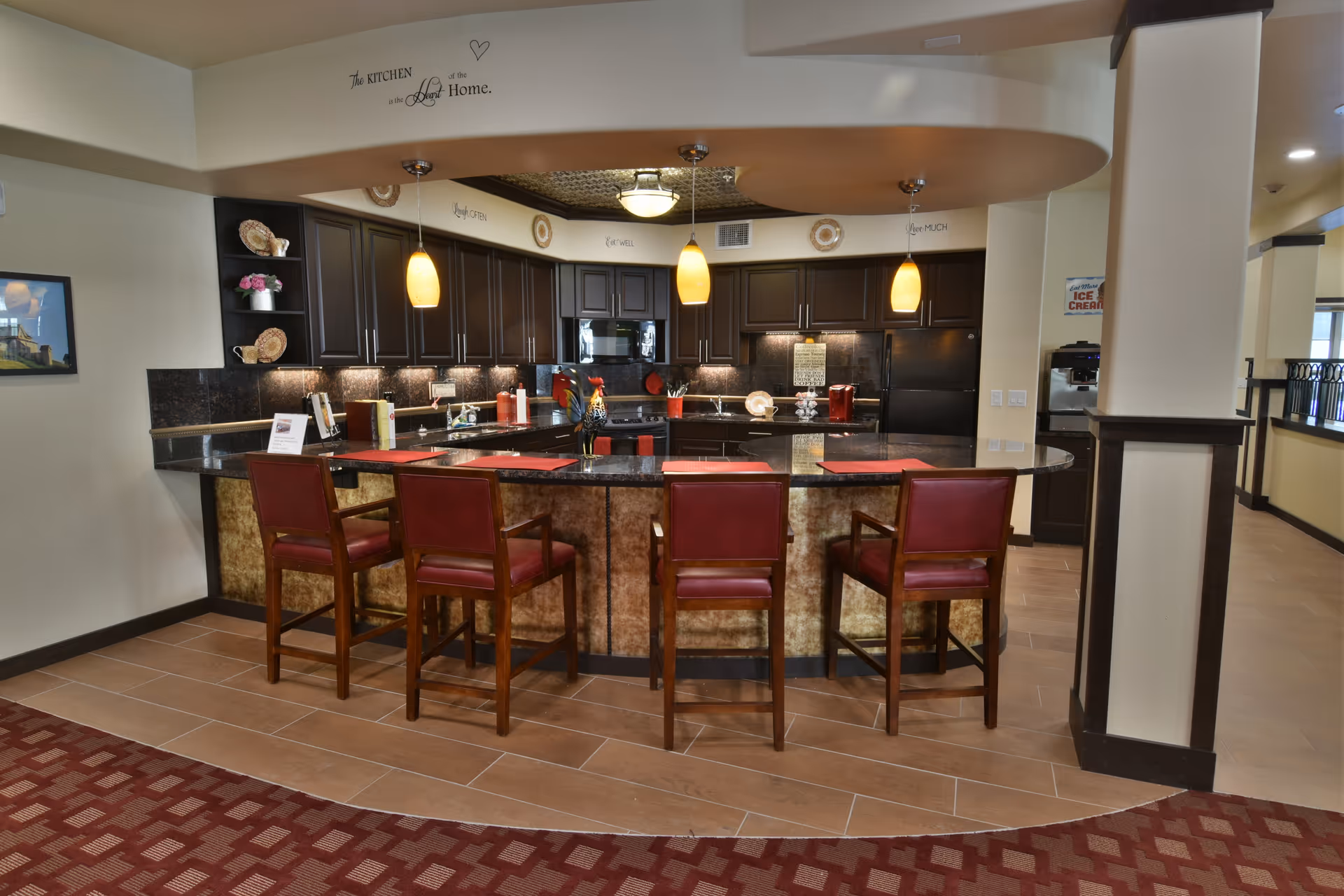 A modern kitchen area with a curved granite countertop island surrounded by four red cushioned wooden bar stools. The kitchen features dark wood cabinets, a black refrigerator, microwave, and stove. Three pendant lights hang above the island, and decorative plates and flowers are displayed on shelves. The wall above the cabinets has inspirational text and a quote that reads, 'The kitchen is the heart of the home.'