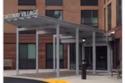 Entrance area of Gateway Village facility showing a covered walkway supported by metal posts leading to glass doors, with brick walls and windows in the background.