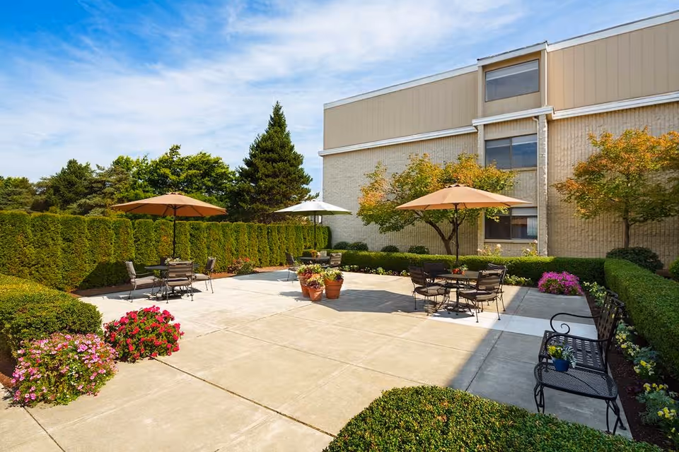 Outdoor patio area at Delaware Plaza Assisted Living Community with several tables and chairs under large umbrellas, surrounded by neatly trimmed hedges, flowering plants, and trees, adjacent to a beige building under a partly cloudy blue sky.