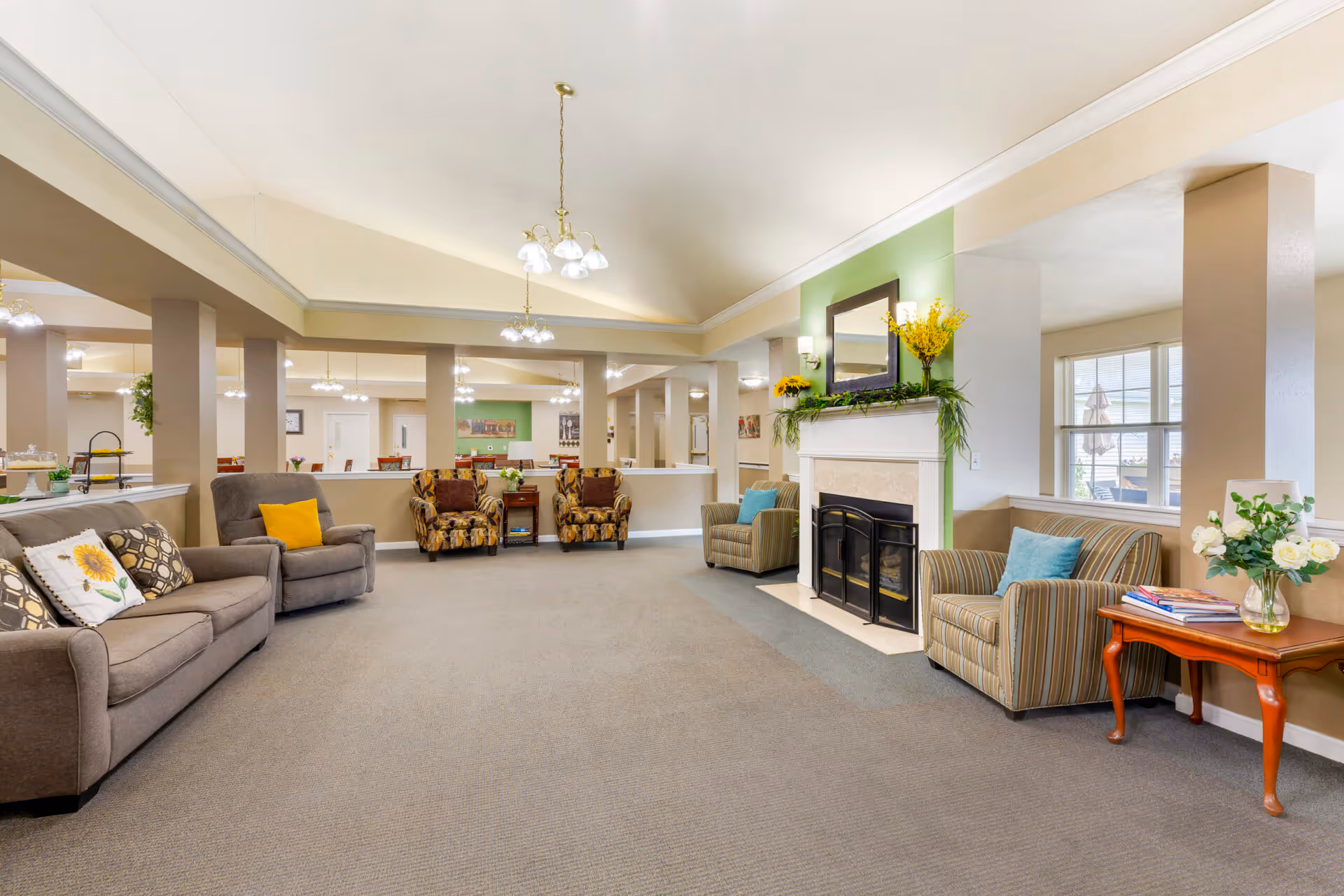 A spacious senior living facility common area with multiple seating options including sofas and armchairs arranged around a fireplace. The room features a high ceiling with hanging light fixtures, a large mirror above the fireplace decorated with yellow flowers and greenery, and windows letting in natural light. There are side tables with books and flower vases, and the overall decor is warm and inviting.