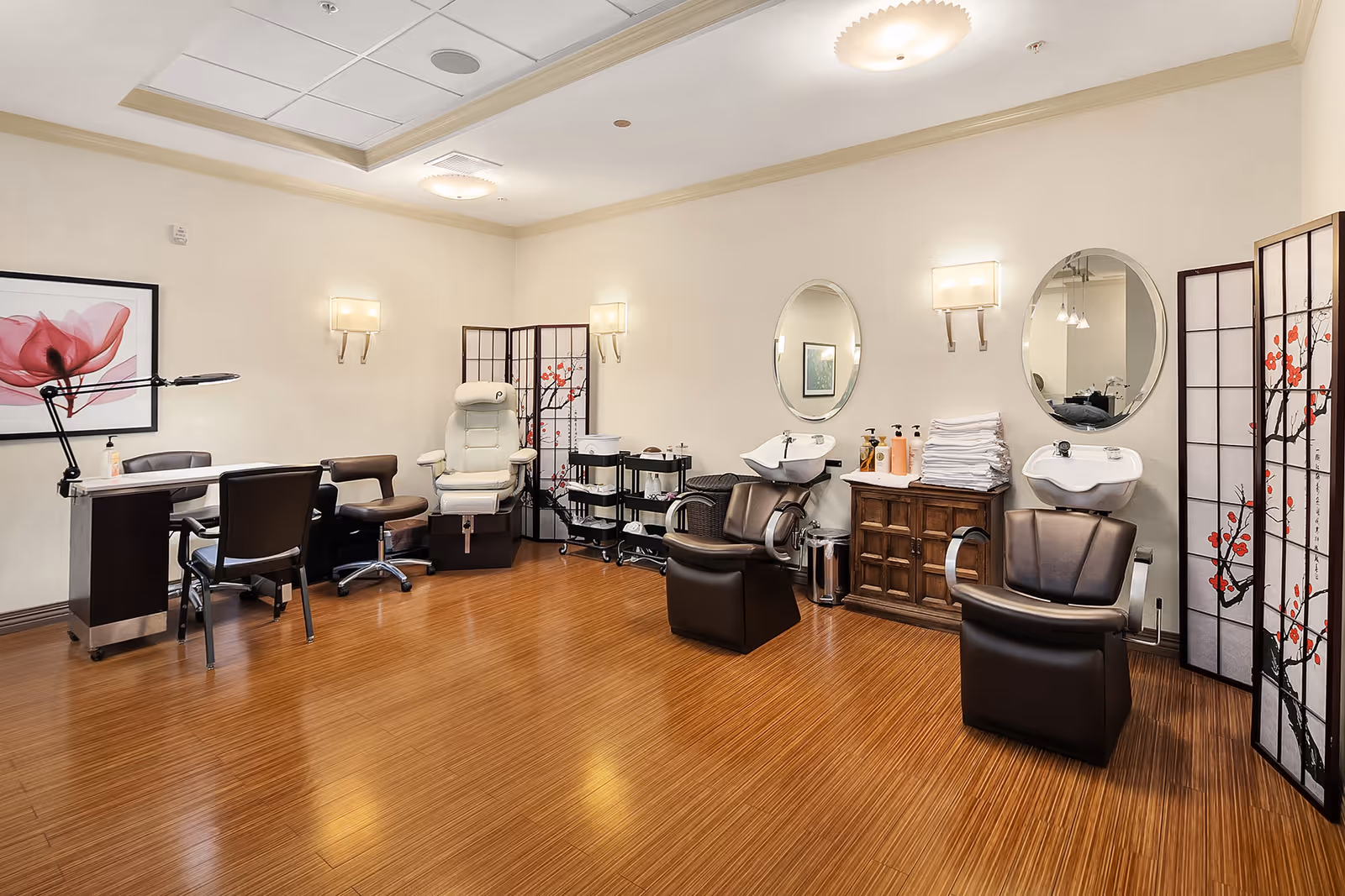 A well-lit salon area with two black salon chairs in front of wash basins and mirrors, a white pedicure chair, a manicure table with chairs, and decorative folding screens with floral designs. The room has wooden flooring and light-colored walls with framed artwork and wall sconces.