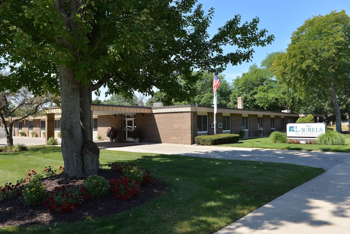 Front entrance of a single-story brick senior living facility with a flag, landscaped lawn, and a sign for The Laurels of Hudsonville.