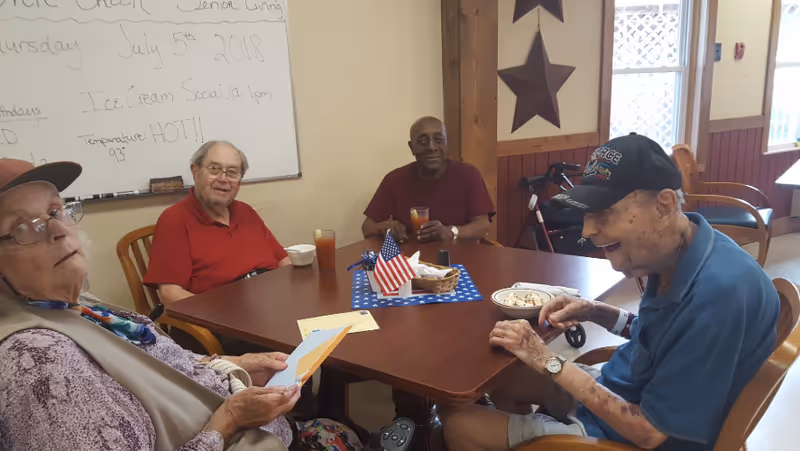 Four elderly individuals sitting around a wooden table in a senior living facility dining area. They are enjoying drinks and snacks, with a small American flag centerpiece on the table. A whiteboard in the background displays the date Thursday, July 5th, 2018, and mentions an Ice Cream Social at 1 pm with a temperature note of HOT 93 degrees.