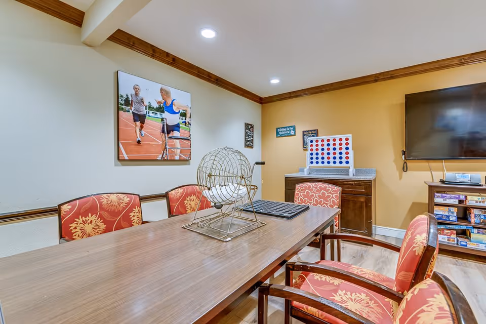 A game room with a wooden table surrounded by red cushioned chairs with floral patterns. On the table is a bingo cage and a large Connect Four game is on a counter against a yellow wall. A flat-screen TV is mounted on the wall above a shelf filled with board games. A photograph of two elderly people running on a track is hung on the adjacent wall.