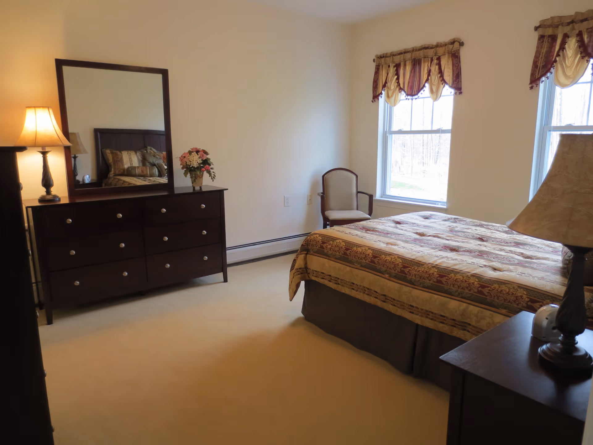 A bedroom with a large bed covered in a patterned bedspread, two windows with decorative valances, a wooden dresser with a mirror, a table lamp, a small flower arrangement, and a single upholstered chair near the window.