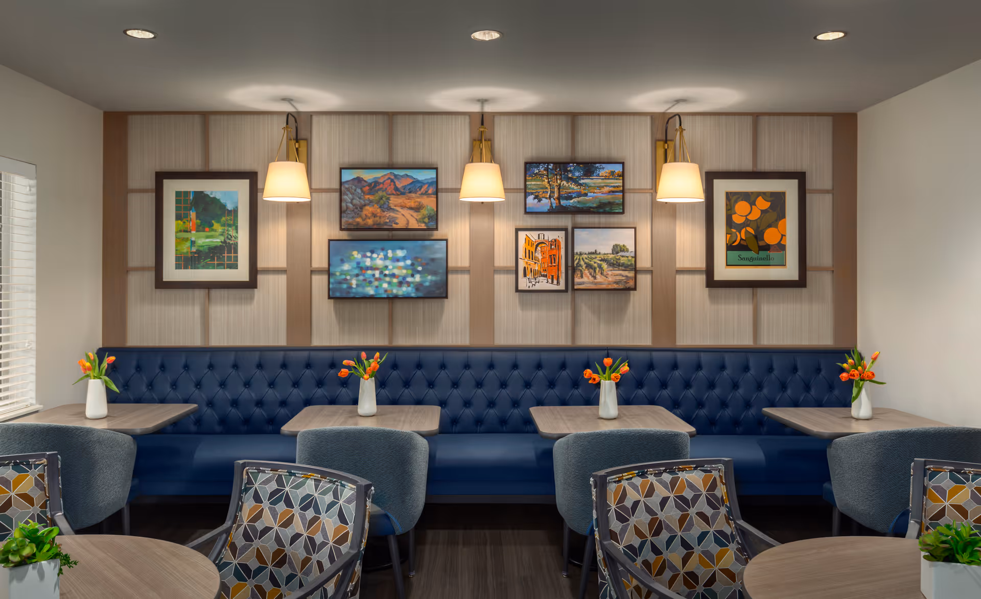 A cozy dining area with a long blue tufted bench along the wall, paired with several small wooden tables and cushioned chairs. Each table has a white vase with orange tulips. The wall behind the bench features a decorative panel with seven framed paintings and three wall-mounted lamps with beige shades.