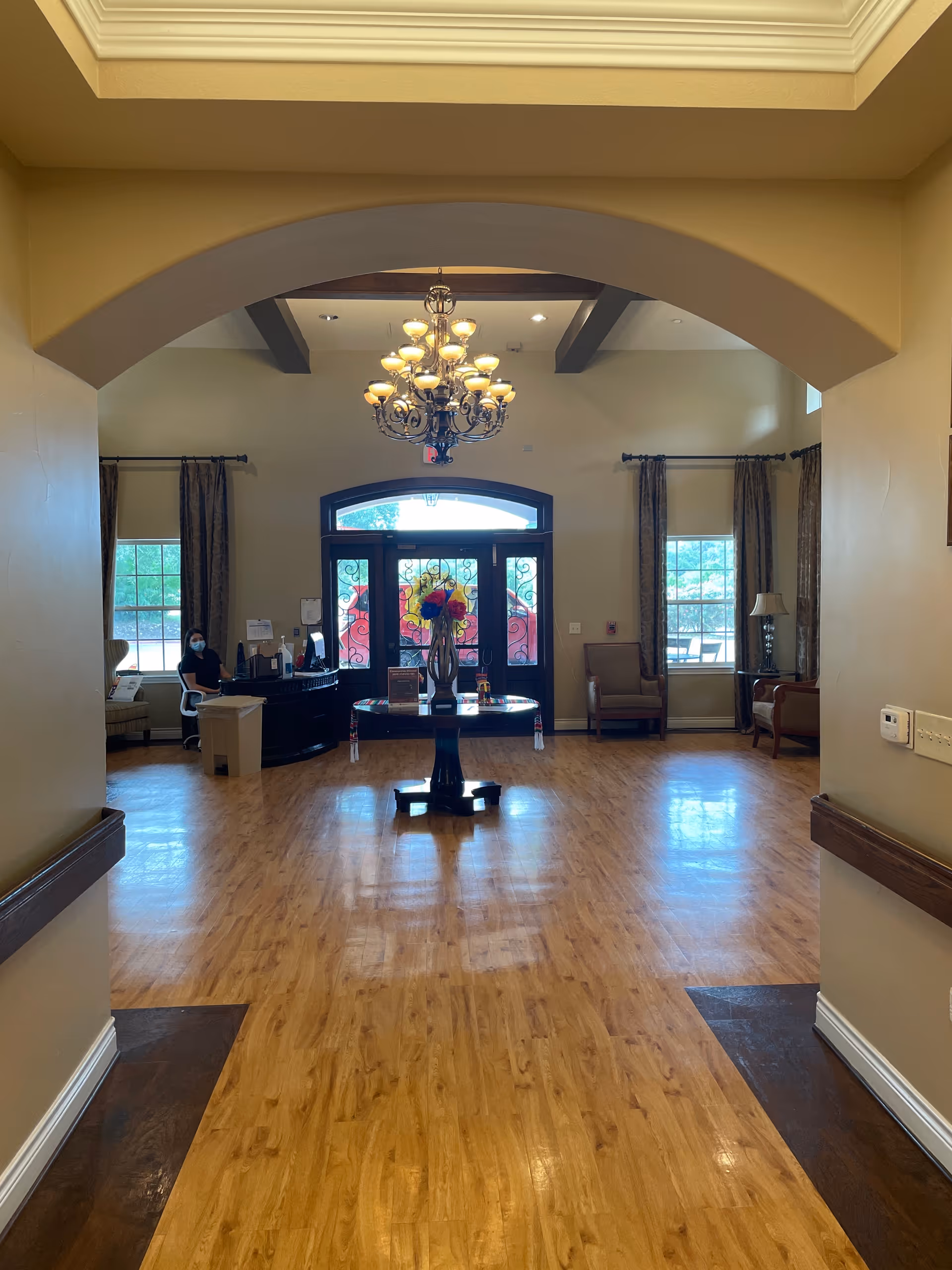 View of a spacious lobby area with wooden flooring, a round table with a flower vase in the center, a chandelier hanging from the ceiling, large windows with curtains, and a reception desk with a person wearing a mask seated behind it. The entrance door with decorative glass panels is visible at the far end.