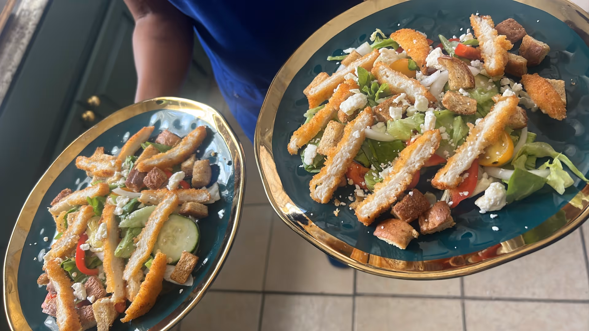 Two plates of salad with lettuce, cucumber slices, cherry tomatoes, croutons, feta cheese, and strips of breaded chicken being held by a person wearing a blue shirt.