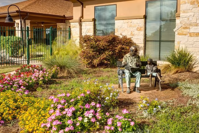Outdoor garden area with colorful flowers and bushes in front of a building with stone and stucco walls. A bronze statue of an elderly man reading a book to a child sitting on a bench is prominently displayed in the garden.