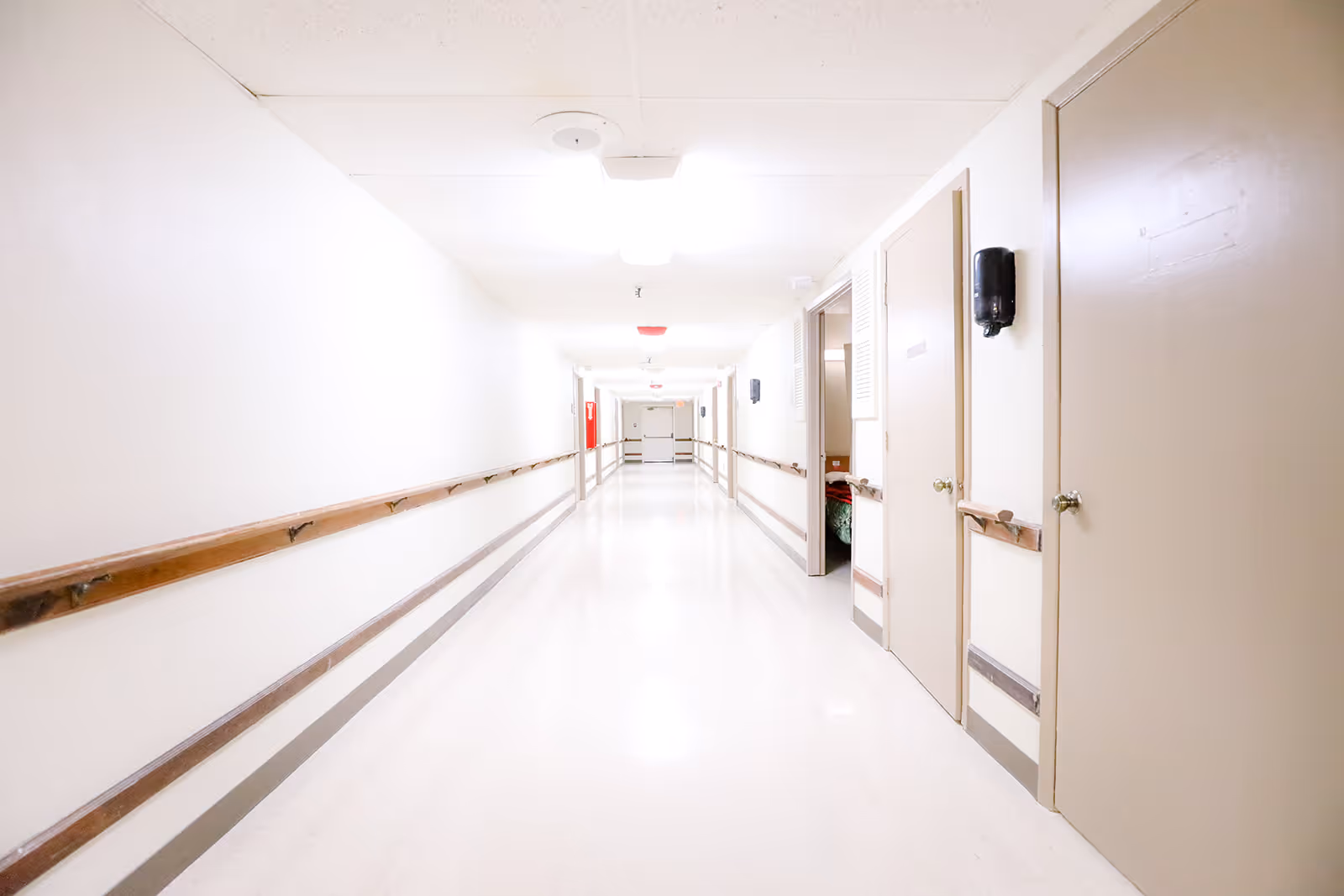 A long, brightly lit hallway in a healthcare facility with white walls, a white floor, and wooden handrails on both sides. Several closed doors line the right side of the hallway, and a fire extinguisher is visible mounted on the left wall in the distance.