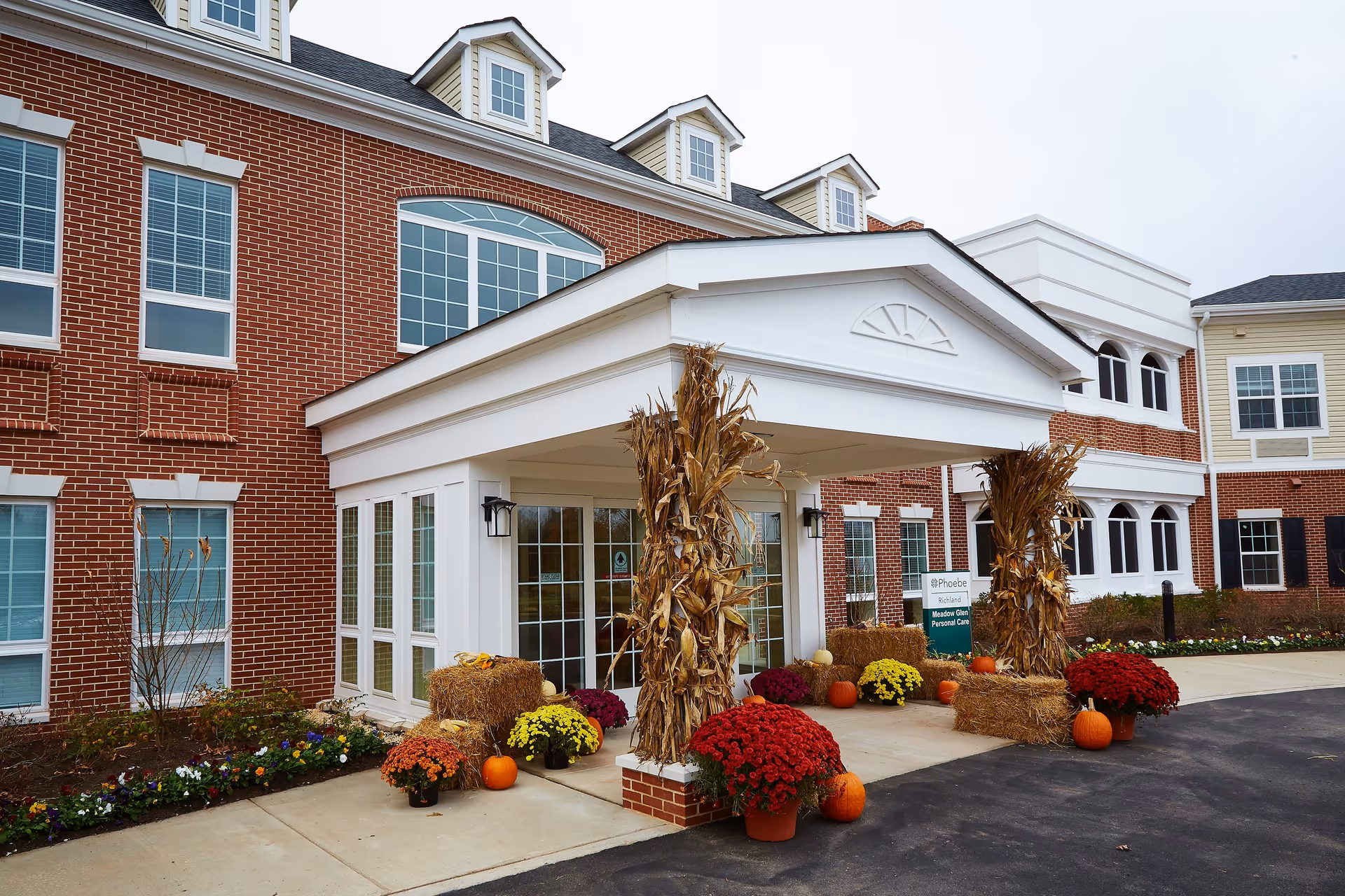 Entrance of a senior living facility named Phoebe Richland decorated with autumn-themed items including hay bales, pumpkins, corn stalks, and colorful potted flowers. The building has red brick walls, white trim, and multiple windows.