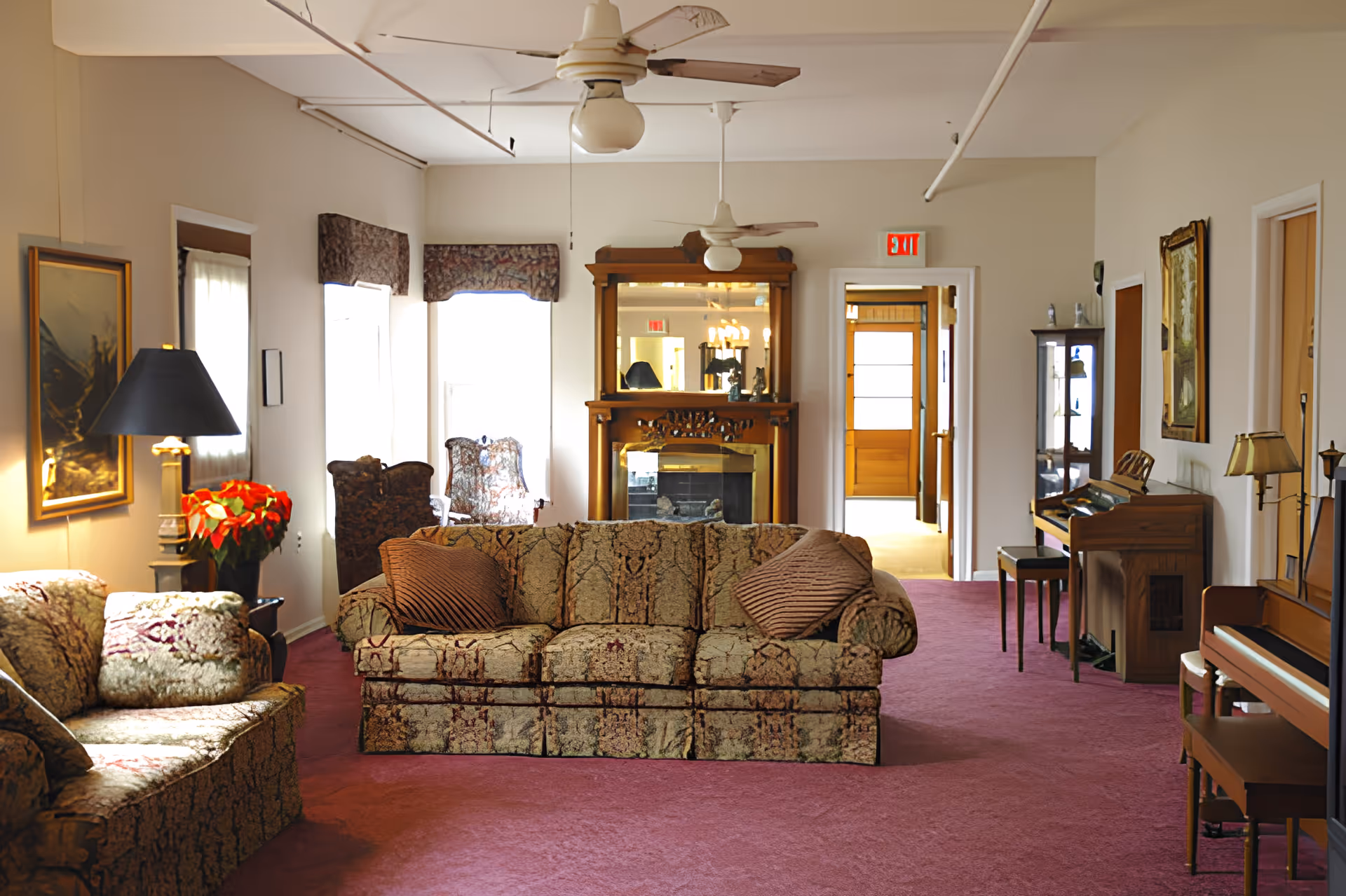 Carpeted common living room with patterned sofas and armchairs facing a fireplace with mirror, piano, and ceiling fans.