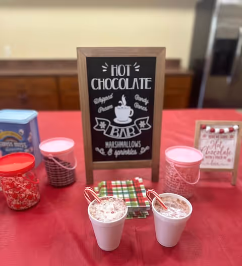 A hot chocolate bar setup on a red tablecloth with two cups of hot chocolate topped with whipped cream and candy canes. There are containers with toppings like marshmallows and chocolate chips, a stack of plaid napkins, and a chalkboard sign that reads 'Hot Chocolate Bar, Whipped Cream, Candy Canes, Marshmallows & sprinkles.'