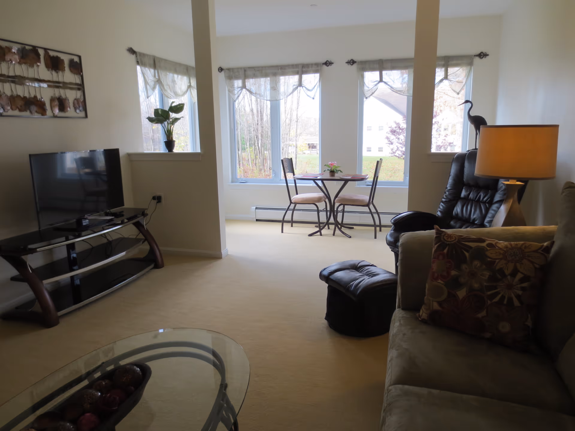 Sunlit living room with a sofa and decorative pillow, TV on a stand, an armchair and lamp, and a small table with two chairs by large windows.