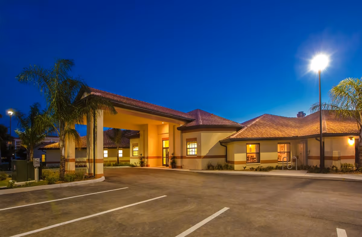 Front entrance of a single-story senior living building lit at dusk with palm trees and an empty parking area.