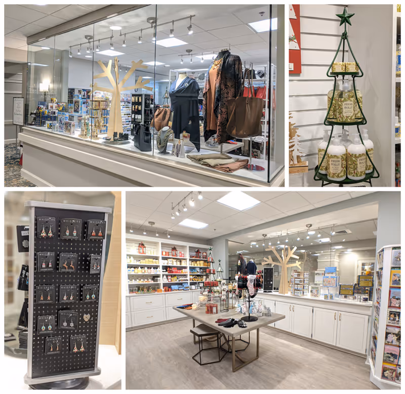Interior view of a gift shop area in Croasdaile Village featuring a display case with clothing and accessories, shelves stocked with various items including lotions and candles, a rotating rack with earrings, and a central table with gloves and other small goods. The space is well-lit with white cabinetry and wood flooring.