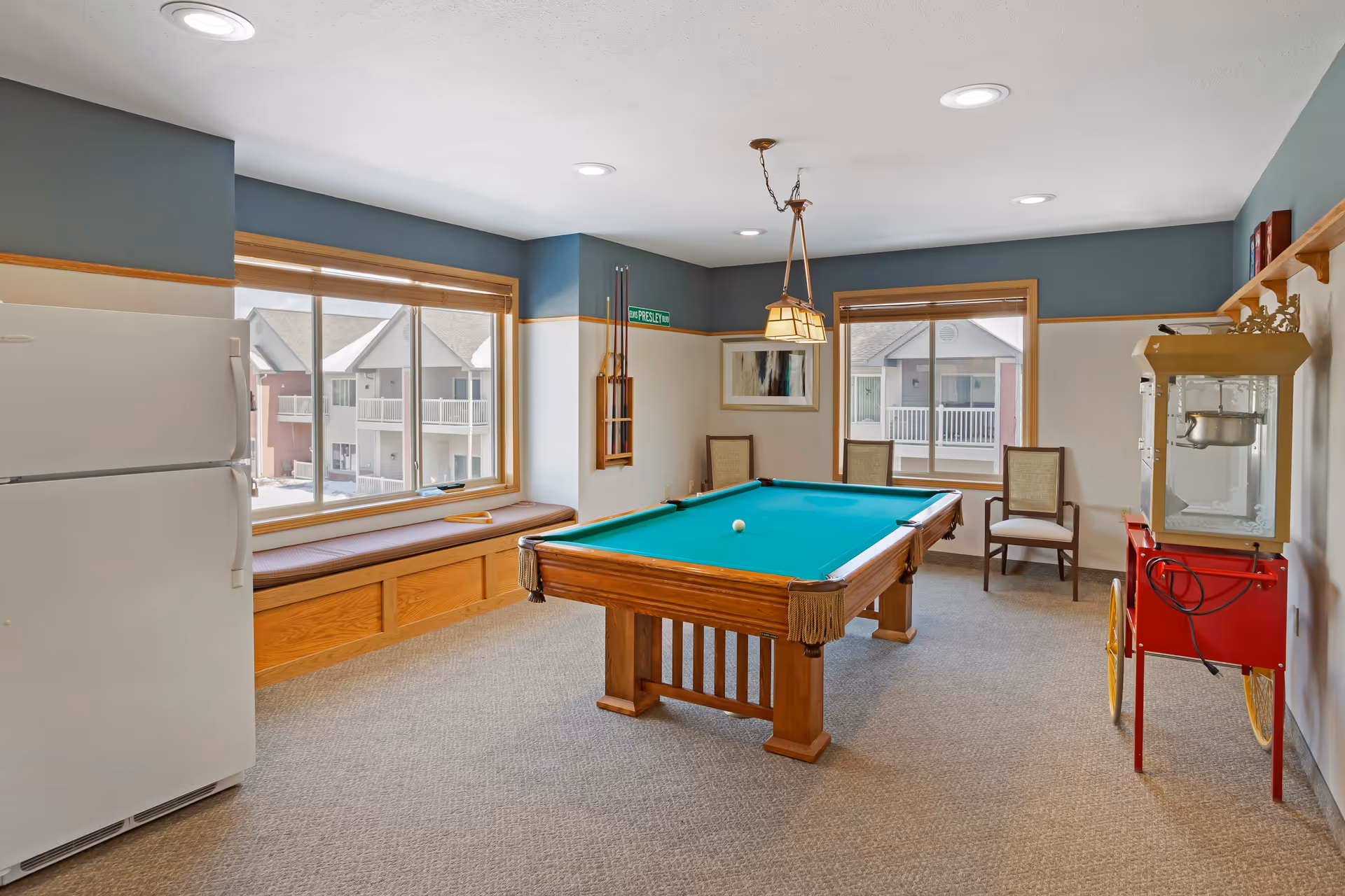 A recreational room with a pool table in the center, a white refrigerator on the left, a popcorn machine on the right, two chairs near the window, and a window seat beneath two large windows showing an exterior view of neighboring buildings.