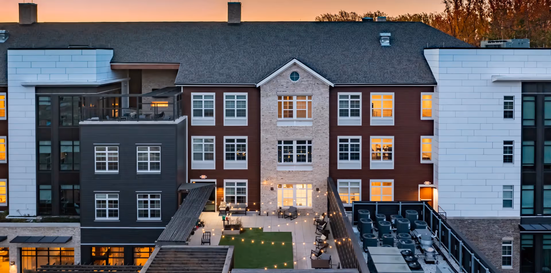 Exterior view of Tribute at Black Hill senior living facility at dusk, showing a multi-story building with lit windows, a rooftop patio with seating, string lights, and surrounding trees with autumn foliage.