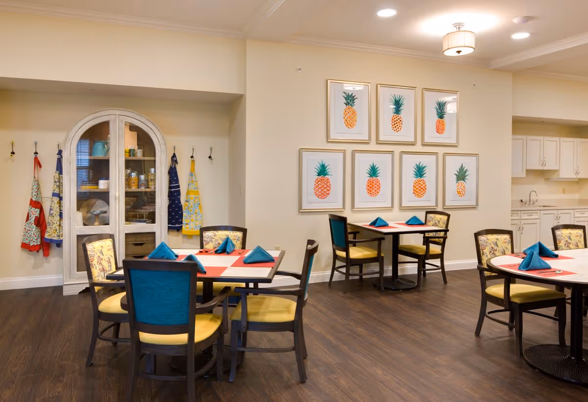 A dining area in a senior living facility with round tables set with red and white tablecloths and blue folded napkins. The chairs have yellow seats and some have blue backs. On the wall, there are seven framed pictures of pineapples arranged in two rows. To the left, aprons hang on hooks next to a white cabinet with glass doors displaying dishes and jars. The floor is dark wood, and the walls are light-colored with ceiling lights providing illumination.