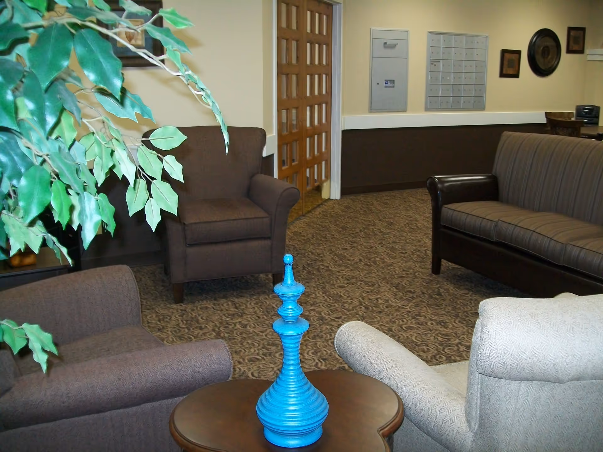 A cozy seating area in a senior living facility with a brown armchair, a striped brown sofa, a light beige armchair, and a small wooden table with a blue decorative object on it. There is a large green plant on the left side and a wall with mailboxes and framed artwork in the background.